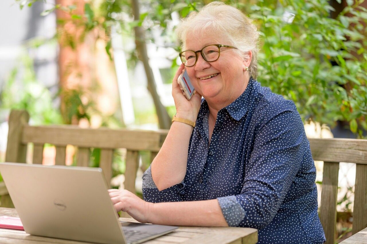 woman in glasses and blue shirt works on laptop at table and makes a phone call